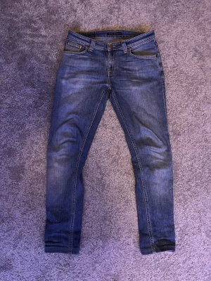 Blå slim fit jeans från Nudie Jeans - Säljer ett par blå slim fit jeans från Nudie Jeans med klassisk femficksmodell och snygga slitningar på framsidan. Jeansen har normal midja, smala ben och är tillverkade i denim med tydlig struktur. Perfekta för dig som gillar en modern och avslappnad stil.