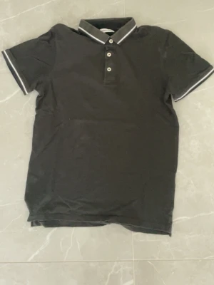 Svart pikétröja med vita detaljer - Svart pikétröja med korta ärmar och vit kant på krage och ärmslut. Tröjan har knappar framtill och klassisk pikékrage. Perfekt för en stilren och enkel look.