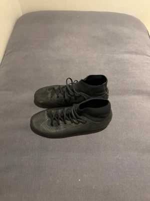 Svarta fotbollsskor med strumpkrage - Säljer ett par helsvarta fotbollsskor med strumpkrage och snörning. Skorna har en modern, slimmad design och är tillverkade i syntetmaterial för bra bollkontroll. Perfekta för dig som vill ha stilrena skor på planen.