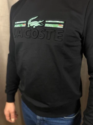 Svart långärmad tröja från Lacoste - Svart långärmad tröja från Lacoste med stort broderat logotyptryck och gröna detaljer framtill. Tröjan har rund halsringning och är tillverkad i mjukt material som känns skönt mot huden. Perfekt för en stilren och sportig look.
