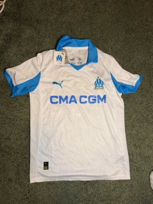 Marseille matchtröja Puma vit/blå - Snygg vit matchtröja från Olympique de Marseille med blå detaljer och klubbmärke på bröstet. Tillverkad av Puma med sponsorlogga CMA CGM framtill och klassisk krage i blått. Perfekt för fotbollsfans som vill sticka ut på planen.