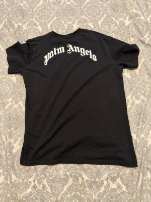 Svart Palm Angels t-shirt - Snygg svart t-shirt från Palm Angels med stort vitt tryck i gotisk stil på ryggen. Klassisk passform och rund halsringning. Perfekt för dig som gillar streetwear och vill sticka ut med en ikonisk logga.