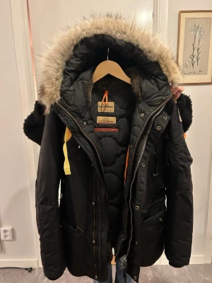 Svart parkasjacka från Parajumpers - Säljer en svart parkasjacka från Parajumpers med stor huva och avtagbar äkta päls. Jackan har flera fickor, gul detalj vid halsen och robust dragkedja. Insidan är quiltad för extra värme och jackan har snygga detaljer i orange och gult.