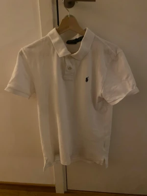 Vit pikétröja från Ralph Lauren - Klassisk vit pikétröja från Ralph Lauren med korta ärmar och krage. Tröjan har två knappar framtill och den ikoniska lilla logotypen broderad på bröstet. Perfekt till jeans eller shorts för en clean och stilren look.