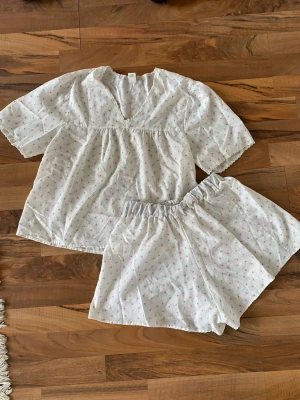 Vit blommig pyjamas med spetsdetaljer - Superfin tvådelad pyjamas från C&A i vit bomull med små rosa blommor och gröna blad. Toppen har v-ringning med spets och knytband, samt korta ärmar. Shortsen har resår i midjan för extra komfort. Perfekt för mysiga hemmakvällar.