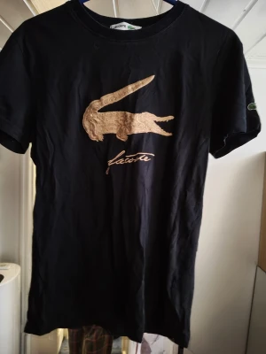 Svart La t-shirt med broderad logga - Snygg svart t-shirt från La med stor broderad guldfärgad krokodil och La-text på bröstet. Klassisk passform och rund halsringning. Perfekt för dig som gillar stilrena plagg med en twist.