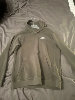 Olivgrön hoodie från Nike - Snygg olivgrön hoodie från Nike med klassisk vit logga broderad på bröstet. Hoodien har en stor känguruficka framtill och en skön huva. Perfekt för en avslappnad och sportig stil.Hör av er om ni har frågor! Priset kan sänkas vid snabb affär!