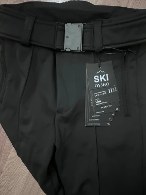 Svarta täckbyxor från Oysho SKI - Svarta täckbyxor från Oysho SKI med flared fit och hög midja. Byxorna har ett brett bälte med metallspänne, dragkedjor vid bensluten och är vattentäta upp till 10K. Perfekta för skidåkning och vinteraktiviteter.