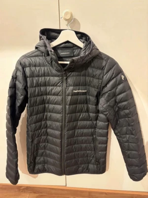 Svart dunjacka från Peak Performance - Snygg svart dunjacka från Peak Performance med huva och dragkedja framtill. Jackan har diskret logga på bröstet och ärmen, quiltad design och två sidofickor med dragkedja. Perfekt för kalla vinterdagar och enkel att matcha med olika outfits.