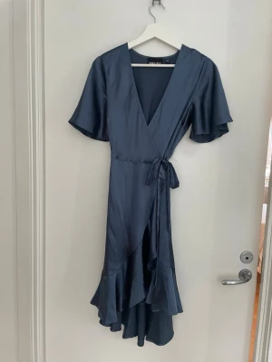 Blå omlottklänning från PIECES - Justerbar midja. Snygg blå omlottklänning från PIECES med v-ringning, volangdetaljer och knyt i midjan. Klänningen har korta, vida ärmar och är tillverkad i ett glansigt material som ger en lyxig känsla. Perfekt för dig som vill ha en elegant och trendig look. Endast använd en gång