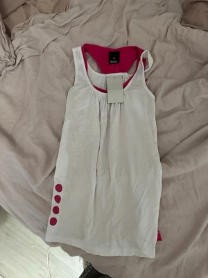 Vit och rosa ärmlös klänning Bench - Snygg ärmlös klänning från Bench i vitt med rosa detaljer och insida. Klänningen har rund halsringning och dekorativa rosa prickar längs ena sidan. Perfekt för en avslappnad och sportig look.