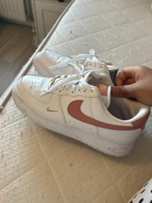 Nike Air Force 1 vita med rosa swoosh - Nike Air Force 1 sneakers i vitt läder med rosa swoosh och guldiga detaljer. Klassisk låg modell med perforerad tå, vita skosnören och Nike Air-logga på plösen och innersulan. Snygg och tidlös design som passar till det mesta.