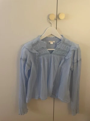 Ljusblå volangblus med spetsdetaljer - Supersöt ljusblå blus med volang vid axlarna och fina spetsdetaljer över bröstet och ärmarna. Blusen har en lös passform, rundad halsringning och är tillverkad i ett tunt, skrynklat tyg som ger en avslappnad känsla.