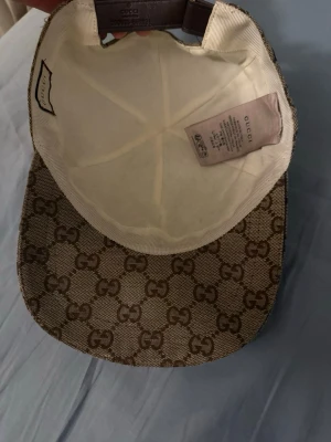 Gucci beige keps  - Säljer en beige keps från Gucci med det klassiska GG-monogrammet över hela kepsen. På sidan finns en grön och röd rand som ger extra lyxig känsla. Kepsen har böjd skärm och justerbart band bak. Materialet är canvas med detaljer i läder.