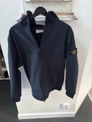 Svart vindjacka från Stone Island - Svart vindjacka från Stone Island med huva och dragkedja framtill. Jackan har klassisk patch med logga på vänster ärm och två fickor framtill. Insidan är fodrad med mjukt material för extra komfort. Perfekt för blåsiga dagar.
