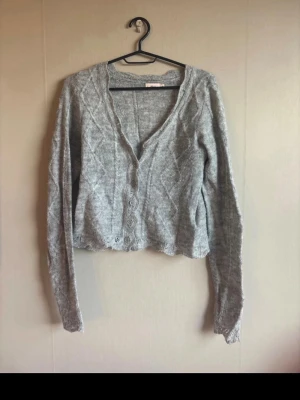 Grå stickad kofta med knappar - Mysig grå stickad kofta med v-ringning och dekorativa knappar framtill. Koftan har ett diskret mönster i stickningen och långa ärmar. Perfekt att slänga på sig över en t-shirt eller linne för en avslappnad look.