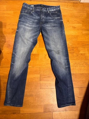 Blå jeans Jack and Jones  - Säljer ett par snygga blå jeans från Jack and Jones med klassisk femficksmodell och raka ben. Jeansen har slitningar och tvättade detaljer för en cool, avslappnad look. Tillverkade i bomull med logga på bakfickan och patch bak i midjan.