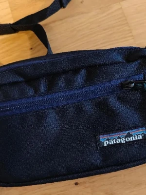 Helt ny mörkblå magväska från Patagonia - Helt ny och oanvänd! Fått denna i present men har redan en som jag använder. 3 fack. Mörk marinblå. För frågor eller fler bilder PM! Snygg mörkblå magväska från Patagonia med justerbart band och smidig plastspänne. Väskan har en dragkedja framtill och är gjord i slitstarkt tyg med mesh på baksidan för extra komfort. 