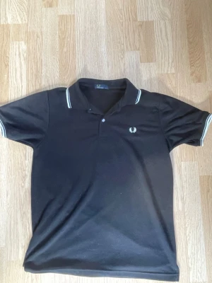 Svart Fred Perry pikétröja - Klassisk svart pikétröja från Fred Perry med vit logga på bröstet och vita ränder på krage och ärmslut. Tröjan har korta ärmar och är tillverkad i ett mjukt bomullsmaterial. Riktigt fet tröja, perfekt till sommaren. 