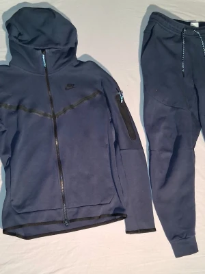 Mörkblå Nike Tech Fleece mjukisbyxor - Säljer mörkblå Nike Tech Fleece byxor med matchande hoodie. Byxorna har dragsko i midjan och ribbade benslut. Materialet är mjukt och stretchigt, perfekt för att träna i eller helt enkelt bara att chilla i på ute eller hemma. Detaljer med svarta Nike-loggor och fickor med dragkedja. Bra pris för allt!