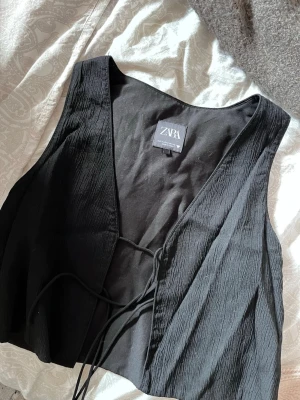 Svart väst med snörning från Zara - Svart linnetopp från Zara, knappt använd med snygg snörning framtill och v-ringad hals. Toppen är ärmlös och har en lätt skrynklig textur