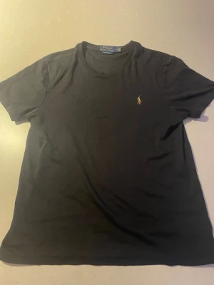 Svart t-shirt från Polo Ralph Lauren - Klassisk svart t-shirt från Polo Ralph Lauren med rund hals och korta ärmar. Ikoniska broderade loggan i gult på bröstet. Skön och enkel modell i mjuk bomull som passar till allt.