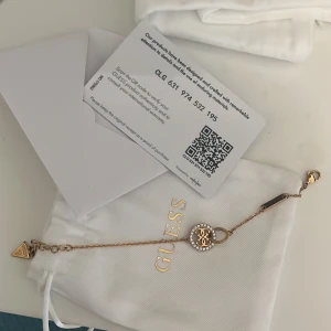 Guess armband - Armband från Guess i roséguld med en rund berlock dekorerad med gnistrande stenar och Guess-logga i mitten. Kedjan är tunn och har en liten trekantig Guess-tag vid låset. Använd vid 1 eller 2 tillfällen, som ny. Medföljer dust bag
