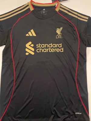 Sprillans Ny Frimpong #30 Liverpool Svart Matchtröja Adidas - Snygg svart Liverpool matchtröja från Adidas med guldiga detaljer och röda sömmar. Framsidan har Liverpool FC-logga och Standard Chartered-tryck i guld. Baksidan har namnet Frimpong och nummer 30 i vitt. Tillverkad i lätt och ventilerande material.