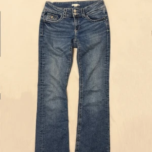 Blå bootcut jeans med fickdetalj - Säljer ett par klassiska blå bootcut jeans med snygga kontrastsömmar och en liten ficka med knapp fram. Jeansen har normal midja och är i ett mjukt denimtyg som sitter skönt. Perfekta till sneakers eller boots.
