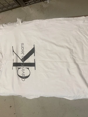 Vit Calvin Klein Jeans t-shirt - Snygg vit t-shirt från Calvin Klein Jeans med stort svart CK-logotyptryck på framsidan. Klassisk rund hals och korta ärmar. Tillverkad i mjukt bomullsmaterial som känns skönt mot huden. Perfekt för en clean och stilren look.