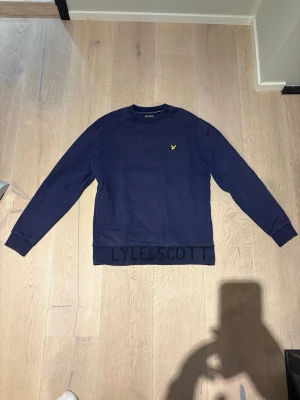 Mörkblå sweatshirt från Lyle & Scott - Mörkblå sweatshirt från Lyle & Scott med relaxed fit. Tröjan har rund hals, långa ärmar och en gul logga broderad på bröstet. Ribbstickade muddar vid hals, ärmslut och nederkant. Perfekt för en chill och stilren look.