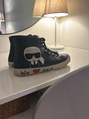 Karl Lagerfeld sneakers svart - Svarta high-top sneakers från Karl Lagerfeld med coolt tryck av Karl-figur med solglasögon på sidan. Texten 'We ♥ karl' finns på sulan. Skorna har snörning och vit sula med svart kant. Perfekt för dig som gillar statement-skor.