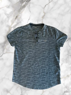  t-shirt Hollister - Snygg gråmelerad henley t-shirt från Hollister med korta ärmar och tre knappar vid halsen. T-shirten har en liten broderad logga på bröstet och är tillverkad i mjuk bomull. Perfekt för en avslappnad och stilren look.