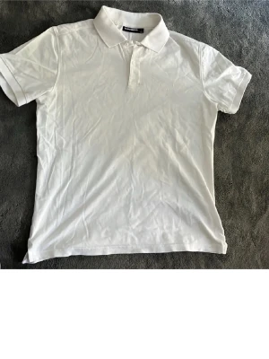 Vit pikétröja från J.Lindeberg - Klassisk vit pikétröja från J.Lindeberg med liten broderad logga på bröstet. Tröjan har krage, korta ärmar och knappar vid halsen. Tillverkad i ett pikématerial som känns svalt och skönt.👕