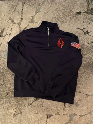 Jeffery Epstein Quarter Zip - Svart sweatshirt med halv dragkedja, röd broderad logga på bröstet och amerikansk flagga på ärmen. Tröjan har hög krage och ribbade muddar. Perfekt för en chill och sportig look.