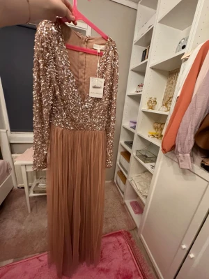 Glittrig långklänning från Maya Deluxe - Långklänning från Maya Deluxe i en champagnefärgad ton. Överdelen är täckt av glittriga paljetter och har v-ringning samt långärmad design. Kjolen är i mjuk, plisserad tyll som ger ett elegant fall. Perfekt för speciella tillfällen.