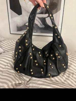 Svart handväska med gulddetaljer - Trendig svart handväska i mjukt material med coola guldfärgade nitar över hela väskan. Väskan har en slouchy form och bred axelrem, perfekt för att ge din outfit en edgy touch. Passar dig som gillar statement-accessoarer och vill sticka ut.