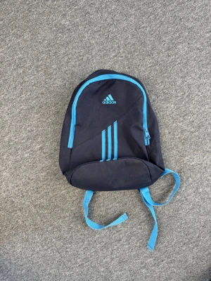 Mörkblå Adidas ryggsäck med blå detaljer - Snygg mörkblå ryggsäck från Adidas med ljusblå detaljer och klassiska ränder på framsidan. Väskan har justerbara axelband, ett stort huvudfack med dragkedja och Adidas-logga framtill. Perfekt för skola eller träning.