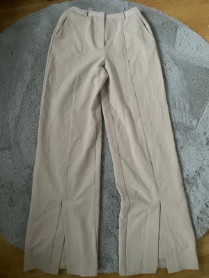 Beige vida kostymbyxor - Snygga beige kostymbyxor med vida ben och pressveck samt slits vid anklarn. Byxorna har hällor för skärp, snedställda fickor fram. Perfekta för en stilren och trendig look.