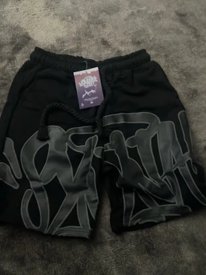 Svarta shorts med graffiti-tryck - Svarta shorts med stort grått graffiti-inspirerat tryck över hela framsidan. De har elastisk midja med snörning och är perfekta för en streetwear-look. Materialet känns mjukt och bekvämt, och shortsen har en avslappnad passform.