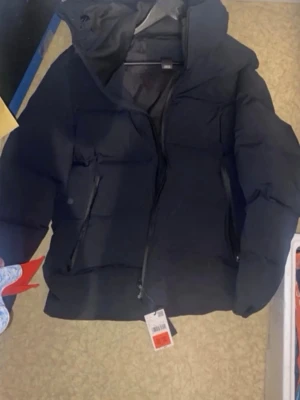 Performance jacka - Snygg svart pufferjacka med huva och dragkedja framtill. Jackan har två snedställda fickor med dragkedja och en quiltad design som ger en modern look. Perfekt för kalla dagar och riktigt varm. Passar dig som vill ha en stilren och enkel vinterjacka.