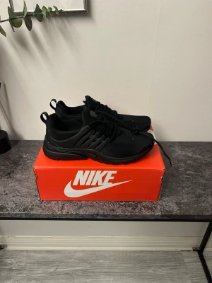 Svarta Nike Air Presto sneakers - Snygga helsvarta Nike Air Presto sneakers med mesh-ovandel och plastdetaljer på sidorna. Skorna har en sportig och stilren design med diskret Nike-logga på sidan och bekväm sula. Perfekta för dig som gillar en clean look och vill ha något som funkar till det mesta.