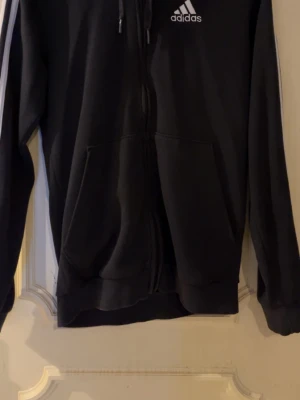 Svart Adidas hoodie med dragkedja - Svart hoodie från Adidas med dragkedja framtill och klassiska vita ränder längs ärmarna. Adidas-logga på bröstet och två fickor fram. Huva med dragsko och ribbade muddar. Perfekt för en sportig och avslappnad stil.