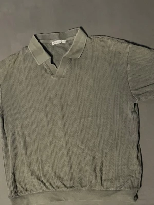 Grön pike från Zara - Säljer en grön piké från Zara med korta ärmar och diskret ribbad struktur. Toppen har klassisk krage och v-ringad öppning framtill. Perfekt för dig som gillar en enkel men stilren look.