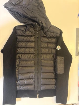 Monclear cardigan - Snygg svart dunjacka från Moncler med quiltad front och huva. Jackan har stickade ärmar och Moncler-logga på armen samt en praktisk ficka på ärmen. Dragkedja framtill och modern, clean look som passar till det mesta. Liten i storlek. Pris går att diskutera. Replic