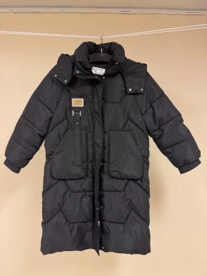 Svart lång vadderad parkas från Fashion - Svart lång parkas från Fashion med huva, stora framfickor och dubbelgående dragkedja. Jackan har quiltat mönster och en snygg detalj med patch på bröstet. Perfekt för kalla vinterdagar och riktigt varm.  100 cm lång. 