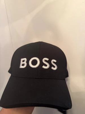 Svart keps från BOSS med brodyr - Snygg svart keps från BOSS med tydlig vit brodyr framtill där det står 'BOSS'. Klassisk böjd skärm och justerbar rem bak. Tillverkad i slitstarkt tyg som ger en clean och modern look. Perfekt accessoar för en streetwear-inspirerad stil.