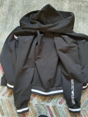 Svart hoodie från Emporio Armani - Snygg svart hoodie från Emporio Armani med vit logga på ärmen och coola vita ränder på muddar och nederkant. Klassisk huva och ribbade detaljer ger en sportig vibe. Perfekt för dig som gillar streetwear och exklusiva märken.