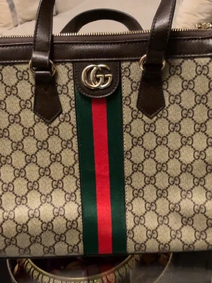Gucci GG Supreme handväska - Snygg handväska från Gucci i beige canvas med klassiskt GG-mönster, mörkbruna läderdetaljer och dubbla handtag. Väskan har en grön och röd rand i mitten samt GG-logga i metall framtill. Perfekt för dig som gillar lyxiga accessoarer med ikonisk design.