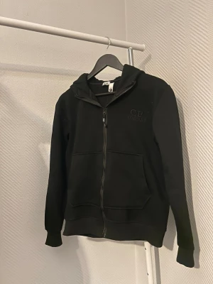 Svart hoodie från C.P. Company - Säljer en svart hoodie från C.P. Company med dragkedja framtill och två stora fickor. Diskret logga broderad på bröstet. Klassisk huva och ribbade muddar vid ärmar och nederkant. Perfekt för en clean och stilren look. Storlek M sitter mer som S